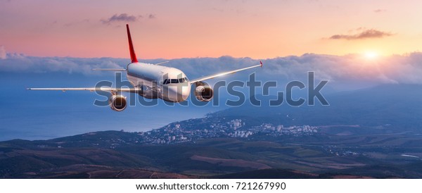 Passenger airplane. Landscape with white airplane\
is flying in the orange sky with clouds over mountains, sea at\
colorful sunset. Passenger aircraft is landing. Commercial plane.\
Private jet. Travel