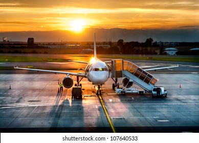 Passenger Airplane On Runway Near The Terminal In An Airport At Sunset Time. Airport Land Crew Doing Flight Service For Passenger Airplane At Sunset Time.