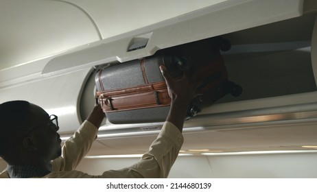 Passenger black man putting luggage on top shelf or cabin compartment on airplane, business man lift suitcases putting overhead locker on airplane, Travel concept