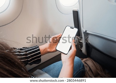 Passenger holding a blank-screen smartphone while seated next to an airplane window, showing in-flight relaxation and mobile connectivity during travel. Casual clothing, tray table and seatback pocket visible.