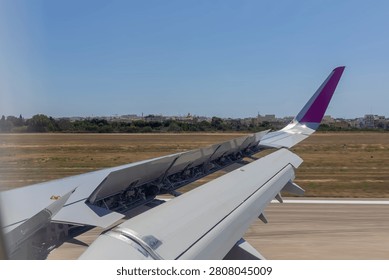 Passenger view capturing the airplane wing with flaps deployed against a clear blue sky, showing airport runway and buildings in the background during the landing process
