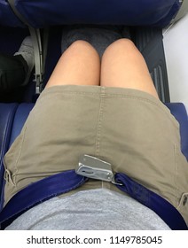 Passenger Who Wears Brown Short Skirt Jeans Is Fastening Blue Seat Belt In Airplane Cabin. There Are Tray Table And Seat Belt For Passenger In Each Chair On The Plane.