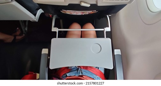 A Passenger Who Wears Short Blue Jeans Is Fastening A Red Seat Belt In Airplane Cabin. There Is Tray Table  Seat Belt For Passenger In Each Chair On The Plane. Girl Wears Pants Has A Tray On The Lap.