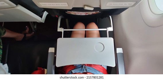 A Passenger Who Wears Short Blue Jeans Is Fastening A Red Seat Belt In Airplane Cabin. There Is Tray Table  Seat Belt For Passenger In Each Chair On The Plane. Girl Wears Pants Has A Tray On The Lap.