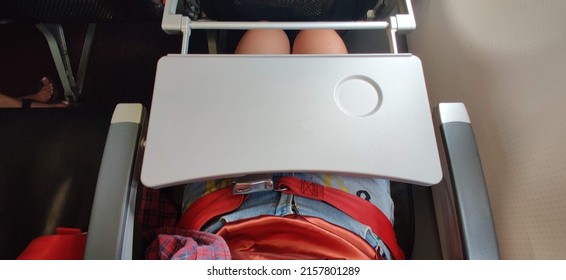 A Passenger Who Wears Short Blue Jeans Is Fastening A Red Seat Belt In Airplane Cabin. There Is Tray Table  Seat Belt For Passenger In Each Chair On The Plane. Girl Wears Pants Has A Tray On The Lap.