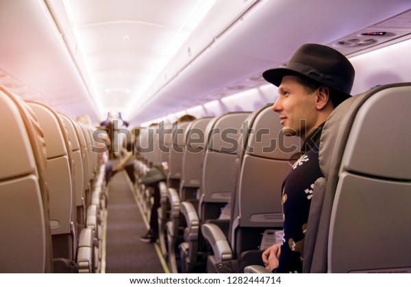 Passengers in an airplane smiling. A young guy holding\
a smartphone is sitting in plane near window with sun rays during\
his business trip .