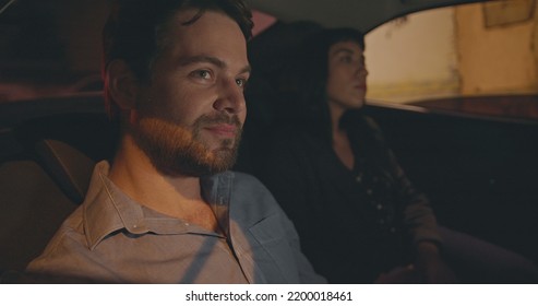 Passengers In Backseat Of Car Riding In City Street At Night. Male And Female Taxi Cab Passenger Commuting After Work