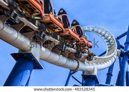 passengers restrained in seats of steel roller coaster trains climbing to a high position in white rail tracks with blue steel supporting posts against blue sky