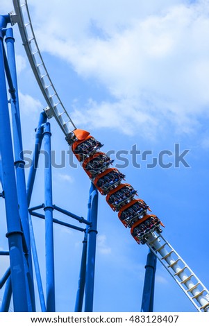 passengers restrained in seats of steel roller coaster trains climbing to a high position in white rail tracks with blue steel supporting posts against blue sky