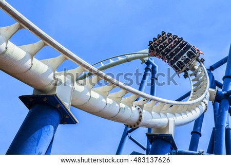 passengers restrained in seats of steel roller coaster trains climbing to a higher position in white rail tracks with blue steel supporting posts against blue sky