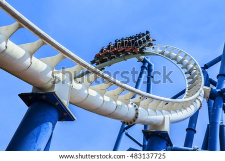 passengers restrained in seats of steel roller coaster trains climbing to a higher position in white rail tracks with blue steel supporting posts against blue sky