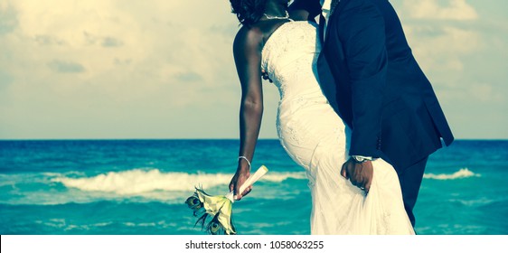 Passionate Afro-American couple kissing on wedding day by the sea. Bride holding bouquet and groom grabbing her leg on the beach. Ceremony union, Valentine Day, love concept. Vintage effect