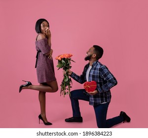 Passionate black man giving his sweetheart bouquet of roses and heart shaped gift for Valentine's Day on pink studio background. Happy woman getting flowers and present from her boyfriend, full length