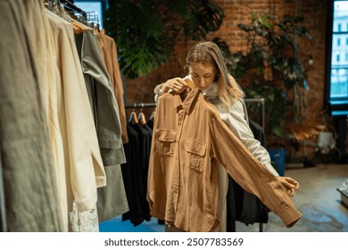 Passionate calm woman trying on clothing items on hanger in apparel shop. Retail therapy for compulsive buyer, interest in selecting garments. Female in second-hand shop, department store, swap meet