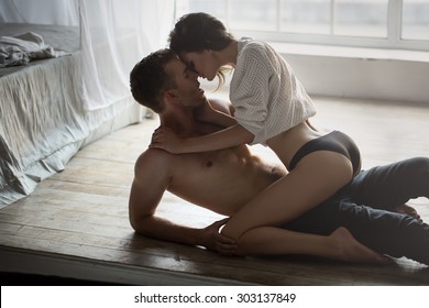 Passionate couple kissing, boy and girl sitting on wooden floor near the rumpled bed opposite the window.