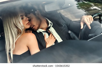 Passionate couple making love in car. Attractive blonde in elegant black dress and handsome businessman in suit are kissing in car.