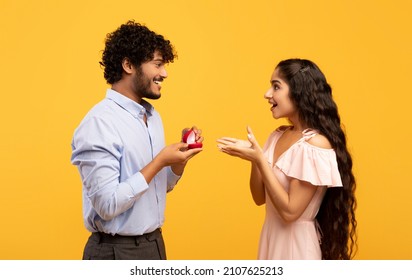 Passionate indian man proposing to his beloved lady, giving her engagement ring on Valentine's Day, yellow studio background. Surprised woman happy to receive marriage offer