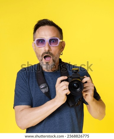 Passionate old photographer beams with excitement, holding mirrorless photo camera, in isolated studio shot on yellow background. Seasoned photographer continues pursue photography passion with zeal