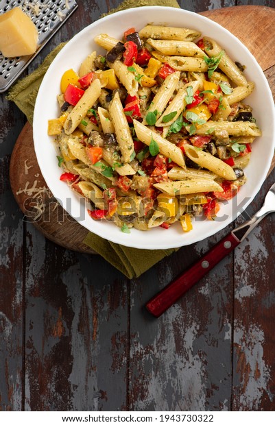 Pasta salad with baked vegetables. Penne pasta with\
baked peppers, eggplant, pesto and cheese in a white plate on a\
dark wooden table top view. Italian food. Rustic style. Copy space\
for text