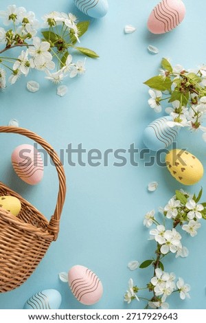 Pastel vertical Easter eggs sit in a woven basket among white blossoms and petals on a soft blue backdrop, creating a festive and welcoming scene for Easter greetings