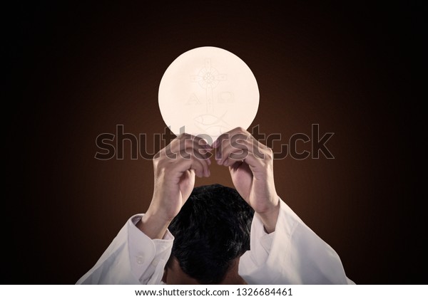 Pastor hands raising a communion bread while\
celebrating a mass on dark\
background