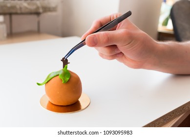 Pastry Chef Adjusting Decoration On An Orange Dessert With Food Tweezers
