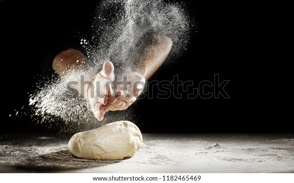 Pastry chef clapping his hands to dust a\
mound of freshly prepared pastry with flour in a freeze motion of a\
cloud of flour midair on black with copy\
space