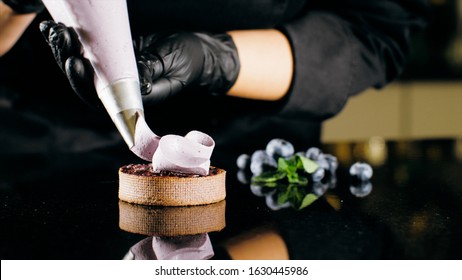 Pastry Chef Decorates Biscuit With Purple Cream From Pastry Bag, Close-up. Preparation Of Blueberry Cake At Commercial Bakery With Piping Bag