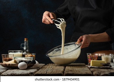 Pastry Chef Kneads A Dough With A Whisk. Backstage Of Cooking Waffle On Rustic Wooden Table With Ingredients On Dark Blue Background. Frozen Motion. Handmade Dessert. Cooking Process.