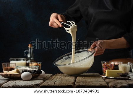 Pastry chef kneads a dough with a whisk. Backstage of cooking waffle on rustic wooden table with ingredients on dark blue background. Frozen motion. Handmade dessert. Cooking process.