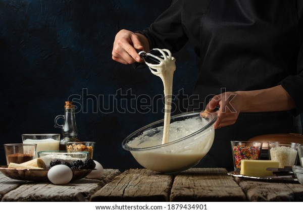 Pastry\
chef kneads a dough with a whisk. Backstage of cooking waffle on\
rustic wooden table with ingredients on dark blue background.\
Frozen motion. Handmade dessert. Cooking\
process.