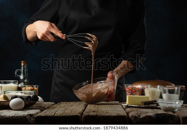 Pastry\
chef testing dough with a whisk. Backstage of cooking waffle on\
rustic wooden table with ingredients on dark blue background.\
Frozen motion. Handmade dessert. Cooking\
process.