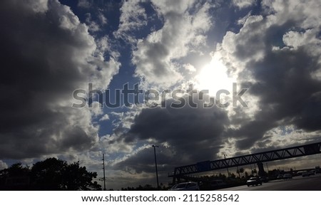 A patch of dark and white clouds stand against a blue sky over a walkover or pedestrian crossing over a busy highway.