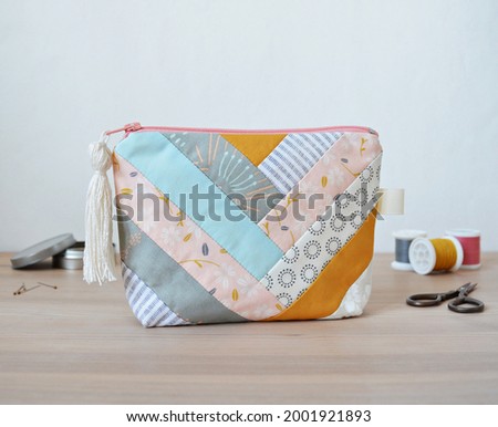 Patchwork zipper pouch with tassel zipper pool, thread spools, little scissors and pins in metal jar on wooden desk over whit
