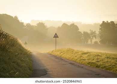 A patchy road winds through the misty countryside, illuminated by a soft morning sunrise. A lone street sign stands beside the road, with a fog-covered open field, and distant hill in the back.