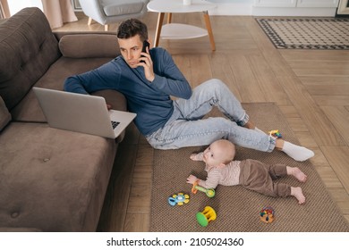 Paternity Leave And Fatherhood. Young Father Freelancer Sitting On Floor, Taking Phone Call, Using Laptop While His Toddler Baby Is Playing Nearby. Busy Dad Working From Home, Solving Family Problems