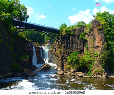 Paterson's Great Falls Historic Park in New Jersey offers a view into the narrow waterfall gorge with a walking bridge above and an American flag flying overhead. 