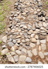 A path made by hand and time. Smooth river stones of grey, tan, and rust set loosely in red earth, winding through dry grass under bright sun. No mortar, no straight lines — just stones placed where they fit. Weeds push through the gaps. Each step crunches. This is how village paths remember footsteps.