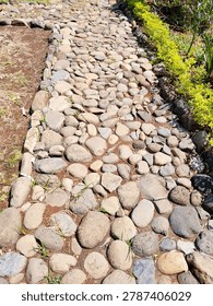 A path made by hand and time. Smooth river stones of grey, tan, and rust set loosely in red earth, winding through dry grass under bright sun. No mortar, no straight lines — just stones placed where they fit. Weeds push through the gaps. Each step crunches. This is how village paths remember footsteps.