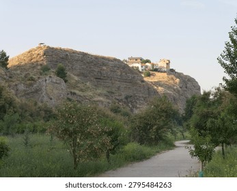 A path runs through green fields leading to a village on a hilltop. The sun shines down on the rocky terrain and trees along the path. People may walk on this day.