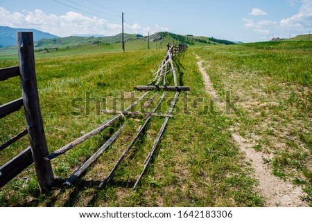Pathway along broken wood fence and poles with wires in mountains in sunny day. Beautiful sunny alpine landscape with footpath along field behind long fence in highlands. Vivid mountain scenery.