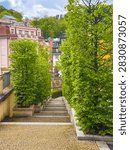 A pathway leads down through trees in Karlovy Vary. Buildings can be seen in the background. It is a clear day with some clouds in the sky.