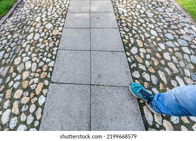 Pathway Made Of Stones With Huge Gray Bricks In The Center, Male Leg In Blue Tennis Shoes Walking On The Footpath, Green Grass, Garden Of A Residential House, Spring Sunny Day