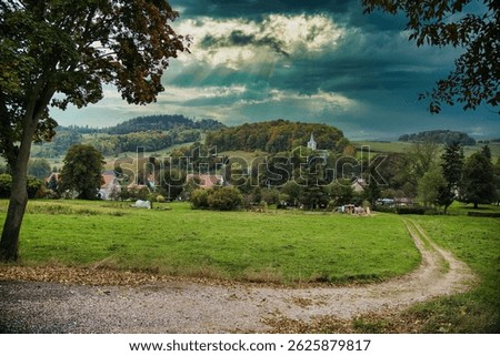 A pathway through a green field to the forest with sun rays coming behind the cloud sin the sky