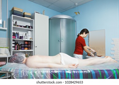 Patient Awaiting Seaweed Body Wrap. Cosmetologist Standing At The Washbasin. At Wrapping Body Is Covered With Active Ingredient And Then Covered With Towel Or Blanket To Create Greenhouse Effect