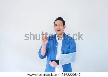 Patient with broken arm in sling, Asian man in casual blue shirt broken hand wearing an arm brace standing over white background with copy space.