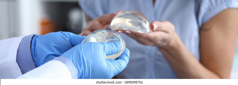 Patient Of Clinic Hold Breast Implant In His Hand And Touch It With Her Finger. Doctor Hold Silicone Material In His Hand And Show Woman.