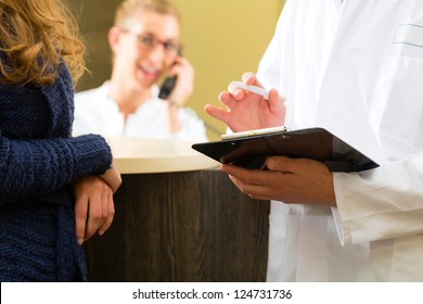 Patient And Doctor In Reception Area Of Office Of Doctor Or Dentist, He Holds A Clipboard, The Receptionist Is On The Phone In Background