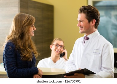 Patient And Doctor In Reception Area Of Office Of Doctor Or Dentist, He Holds A Clipboard, The Receptionist Is On The Phone In Background