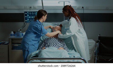 Patient Going Unconscious In Hospital Ward After Surgery At Medical Facility. Nurse And African American Doctor Helping Sick Old Man At Reanimation Room With Oxygen Tube And IV Drip Bag